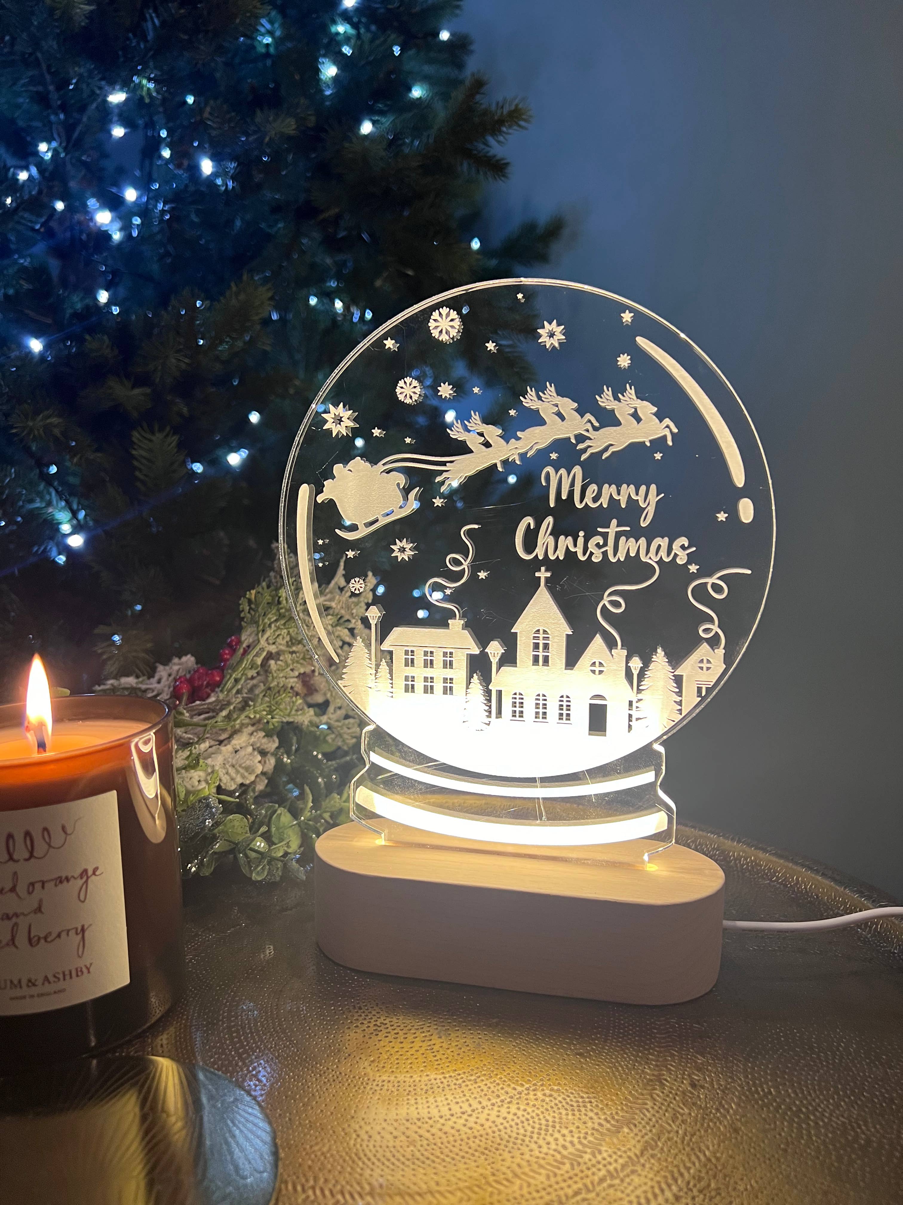A clear acrylic snowglobe with a 'Merry Christmas' message, illuminated from within, placed on a table with a blurred Christmas tree and gifts in the background.